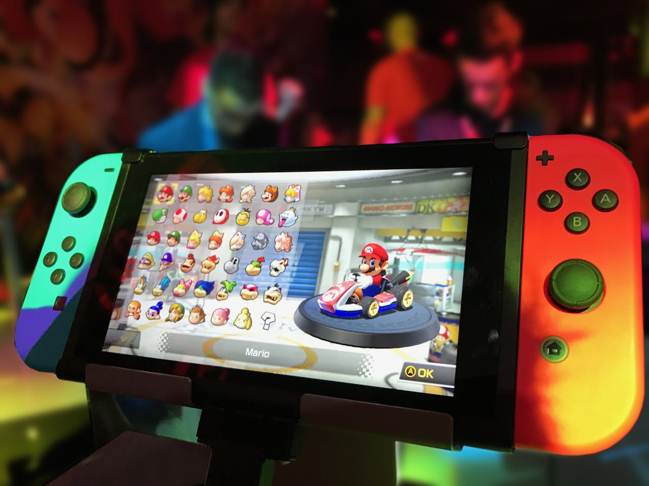 our-mission Close-up of a Nintendo Switch showing Mario Kart selection screen, gaming atmosphere in the background.